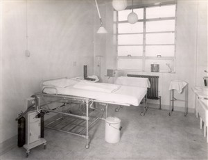 Photo:A delivery table, a gas and air machine, a sphygmomanometer and a drip stand ready for use. A dressing drum and cheatle forceps can be seen on the window ledge and there is an adjustable light over the delivery table, 1951 (Catalogue reference: SBHH-PG-48).