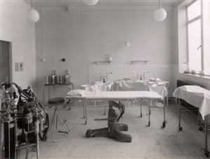 Photo:An operating theatre equipped with an operating table, an anaesthetic machine, instrument tables, prepared sterile drums and a trolley, 1951 (Catalogue reference: SBHH-PG-49).